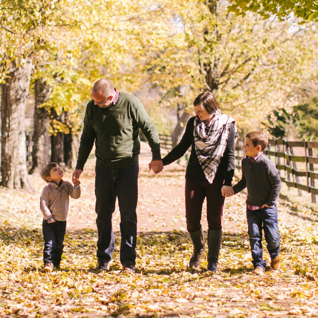 The Magness Family | An image of a mother and father holding hands and walking with their two young boys in a park.