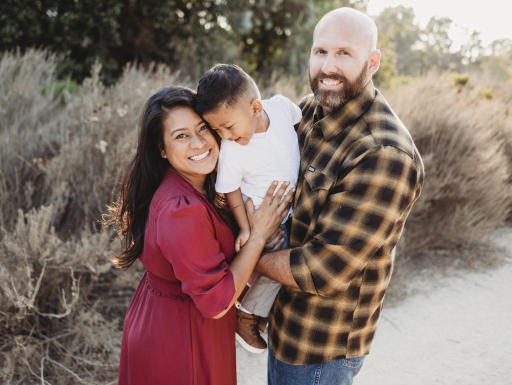 An image of a mother and father holding their male child and smiling in a park