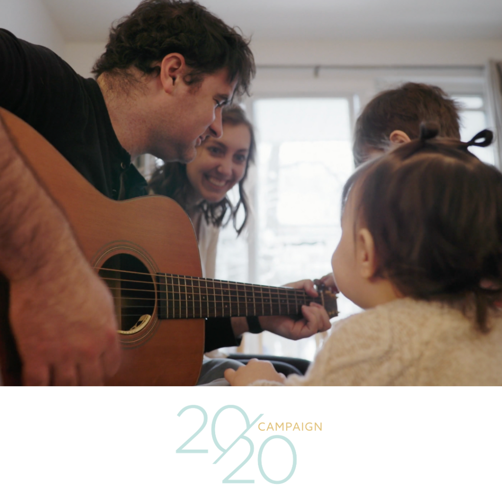 Man playing guitar and family gathered around smiling